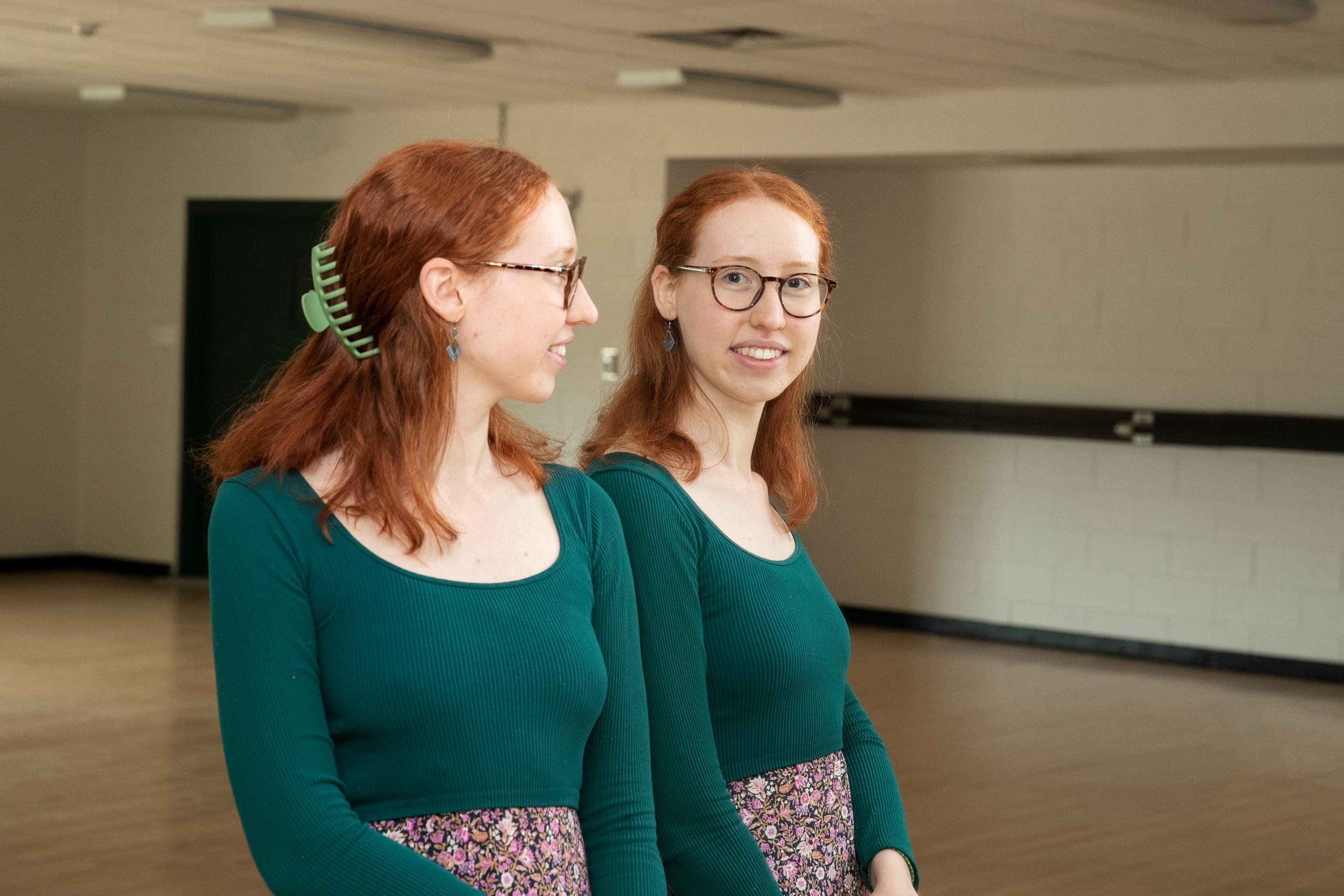 Liz+Bracht+%2724+smiles+into+a+mirror+in+a+dance+studio.+She+is+wearing+a+blue%2C+long+sleeved+top+and+a+floral+skirt.